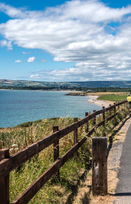 A scenic view of a lush green trail flanked by dense trees, with misty mountains in the background, and a cyclist riding on the Waterford Greenway path.